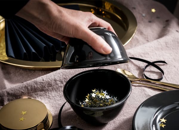 A hand tipping golden stars into a black bowl on a table set with golden and black decorations, plates, trays and cutlery.