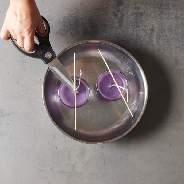 A hand trims the wicks of two purple candles cooling in glass holders, in a tranquil DIY setting reminiscent of an IKEA-style candle project.