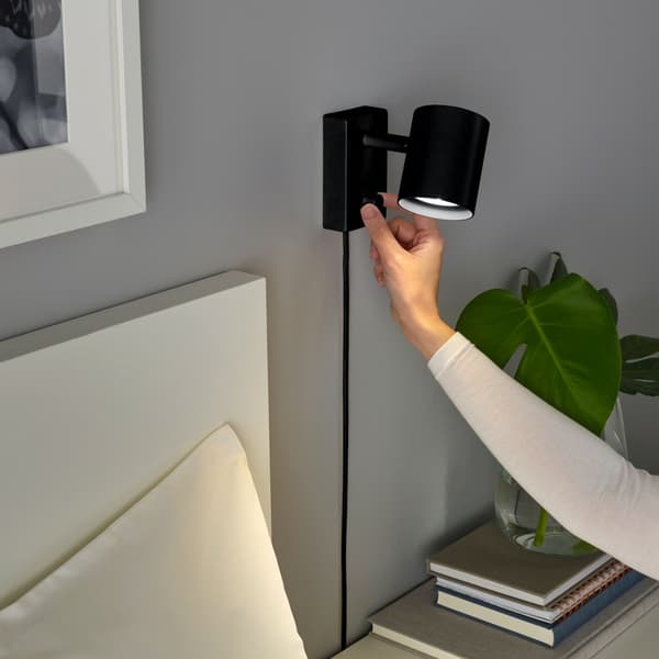 A hand turns on the NYMÅNE wall reading lamp in black colour.