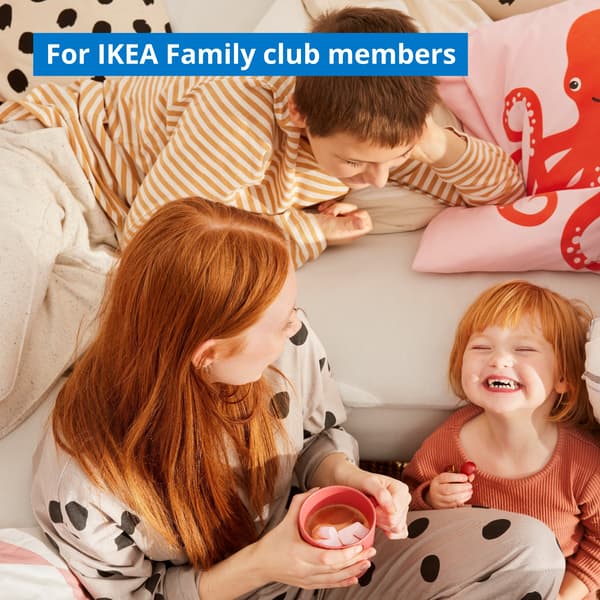 A happy family gathered in the living room by a corner sofa: a red-haired mother, holding a mug of cocoa, watches her little daughter giggle, while the older brother looks on, stretched out on the sofa.
