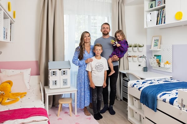 A happy family of four—mother, father, son, and daughter—standing together in a brightly lit, modern children's bedroom featuring white furniture, bunk beds, and a dollhouse.