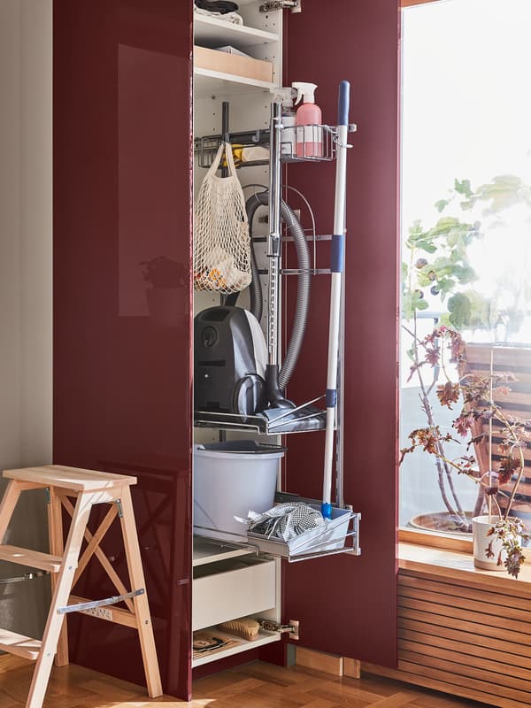 A high cabinet with an open dark red-brown door has a cleaning interior fitting inside holding cleaning utensils.