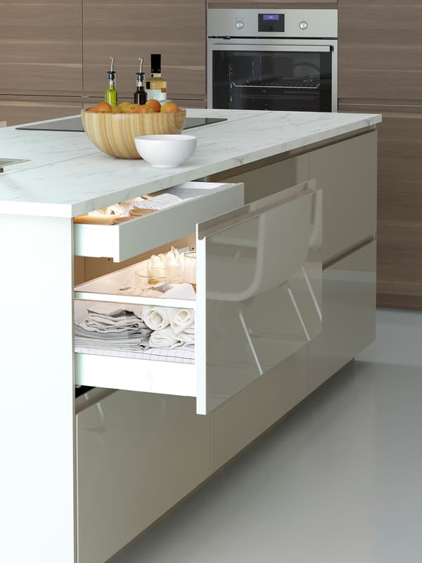 A high-gloss light-beige kitchen island with one open drawer, showing an inner drawer and things such as table linen.