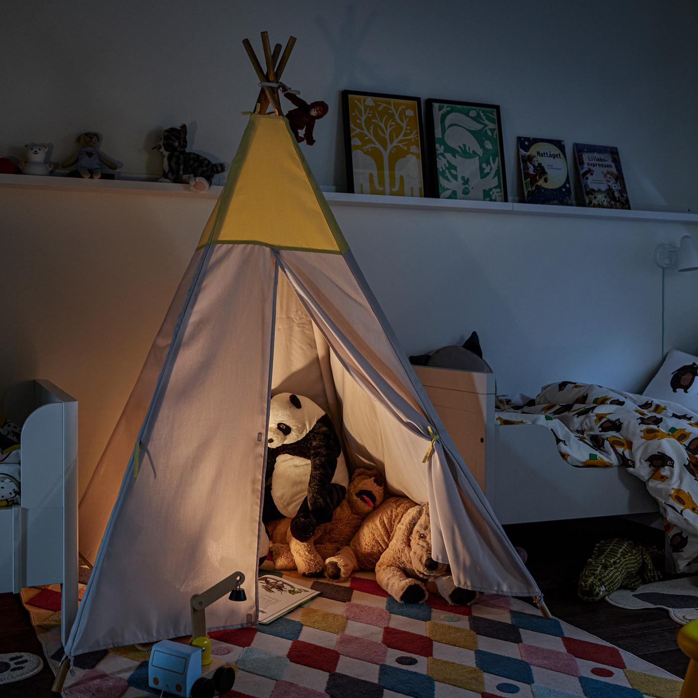 A HÖVLIG tent with various animal soft toys and a lamp inside, in a darkened room between two BUSUNGE extendable beds.