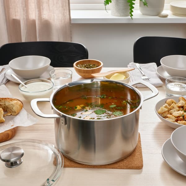A hot pot containing cooked vegetables is standing on a cork board on a set dinner table. 