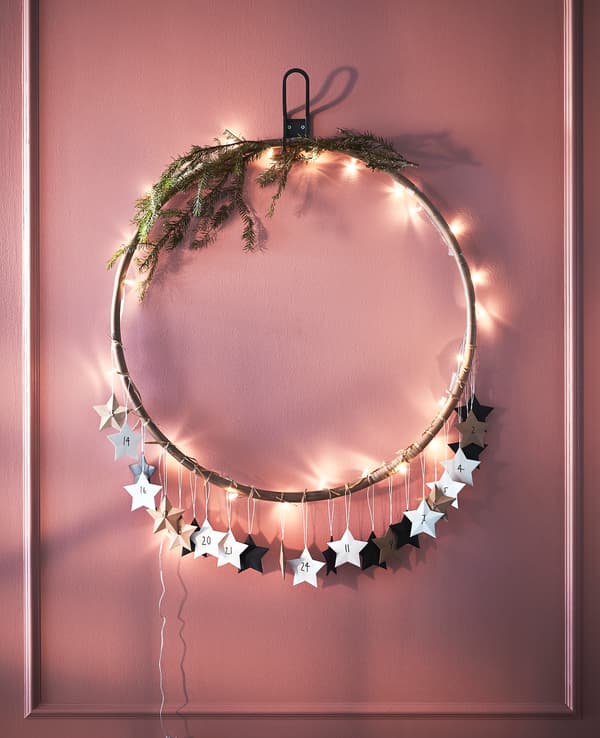 A hula hoop decorated with lights, greenery and paper stars hangs from a wall.