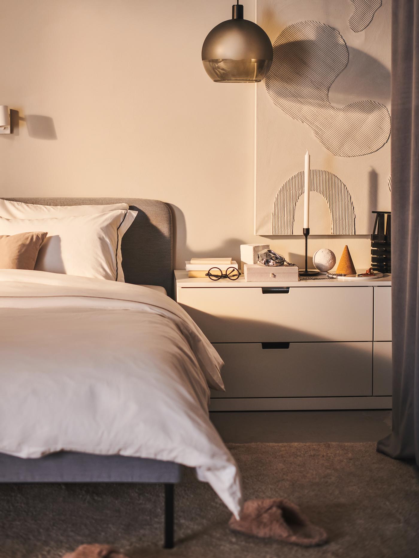 A JAKOBSBYN pendant lamp hangs above a white chest of drawers next to a light grey SLATTUM upholstered bed.