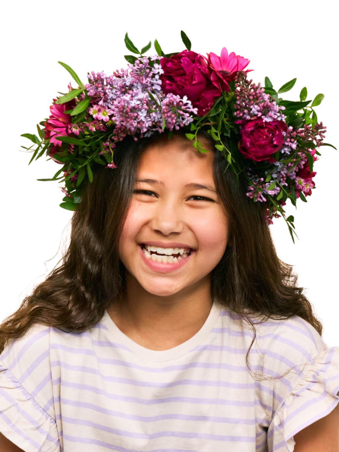 A joyful young girl with long dark hair smiles brightly, adorned with a vibrant floral crown of pink and purple blossoms, celebrating the IKEA Food Festival.