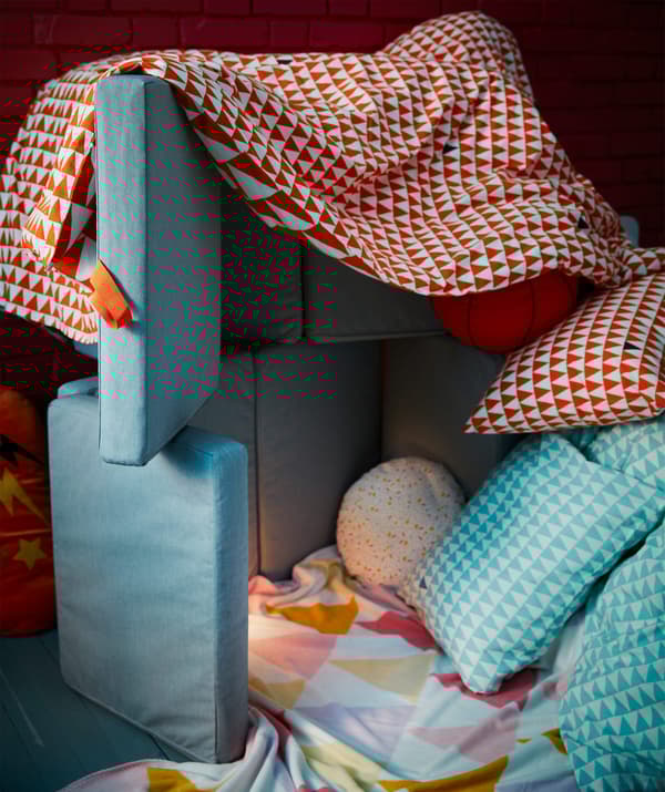 A jumble of bedsheets, cushions, soft toys and a foldable mattress created as a children’s fort.