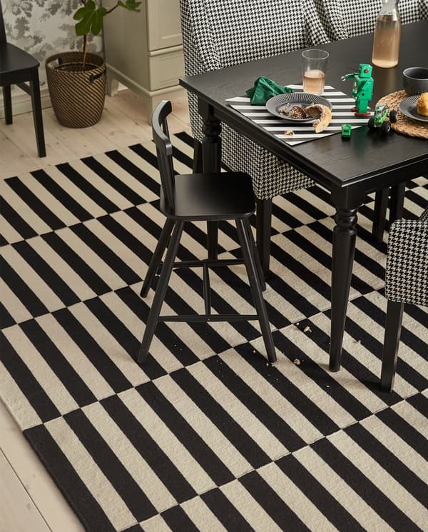 A junior chair stands at a table. Below lies a black/white striped STOCKHOLM low pile rug with some crumbs from a cake.