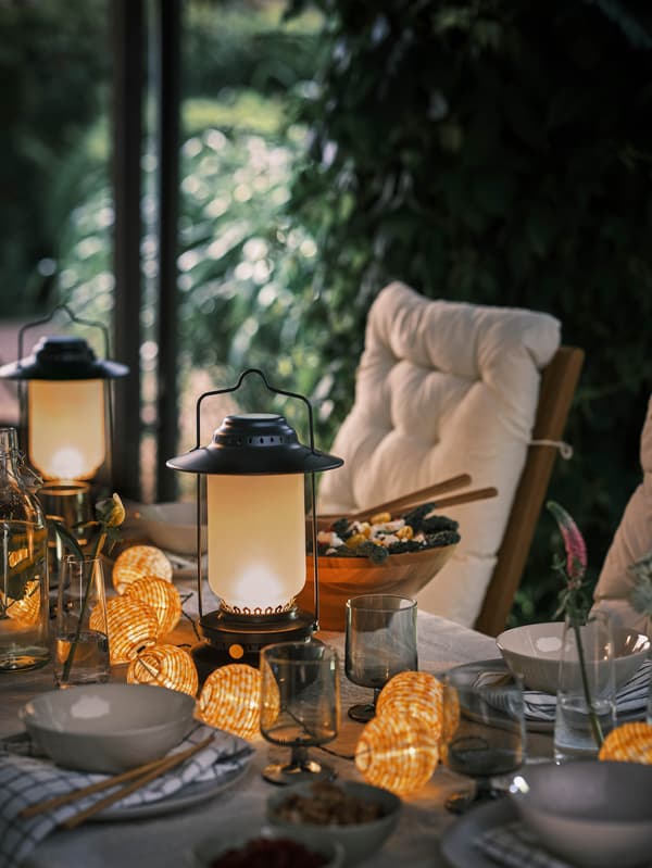 A kertben álló megterített asztalon egy SOMMARLÅNKE világítófüzér és néhány LED-es asztali lámpa. 