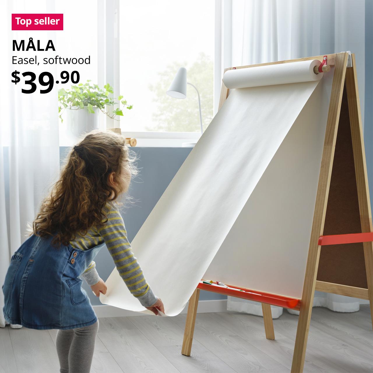 A kid pulling a new piece of canvas on the MÅLA easel board. A reddish pink "Top seller" tag is displayed on the top left corner of the image, followed by a black text overlay image that reads, "MÅLA, Easel, softwood, $39.90"