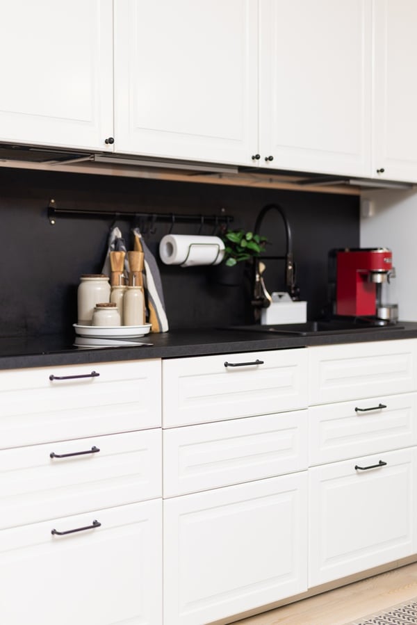 A kitchen countertop with a coffee machine, kettle, and other appliances against a black backsplash, with white drawers below.