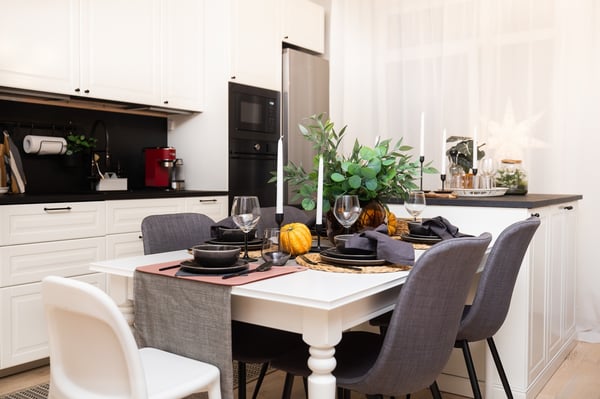 A kitchen dining area with a white table, chairs, and black table setting; the table is decorated with tableware, decorative pumpkins, and green plants, with a modern kitchen in the background.