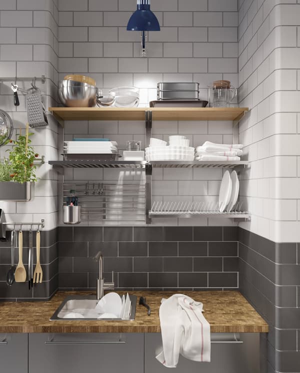 A kitchen in stainless steel, a wood worktop, a blue work lamp and a dish drainer, rails and shelves that are wall-mounted.
