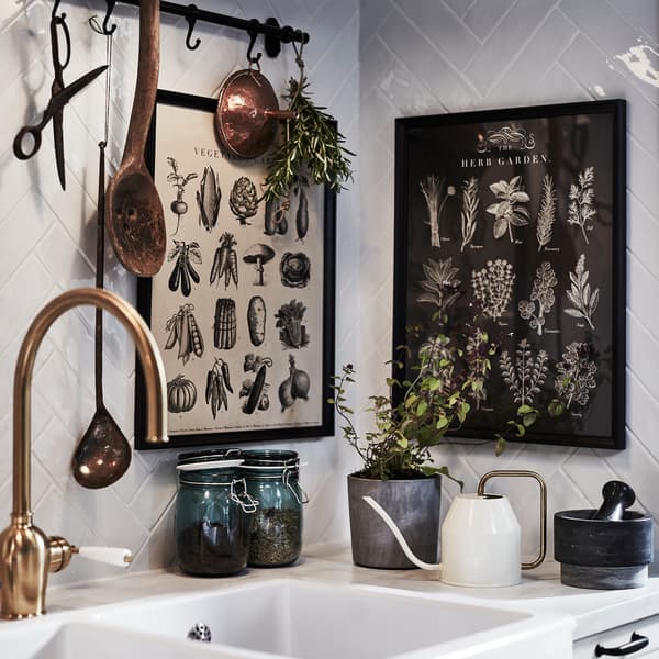 A kitchen sink, a brass-coloured mixer tap, picture frames with herb illustrations, herbs and a brass/white watering can.