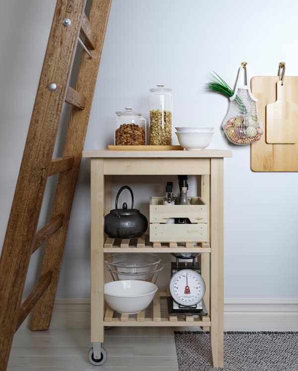 A kitchen trolley in birch stands under a wooden ladder. Kitchen utensils, jars and a teapot are on the trolley shelves.