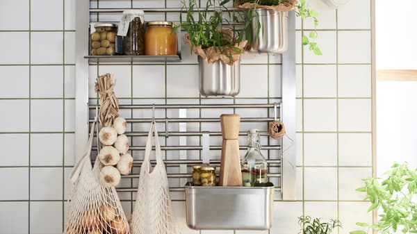 A kitchen wall with a KUNGSFORS wall grid with shelf, hooks and containers, all in stainless steel, holding jars and herbs.