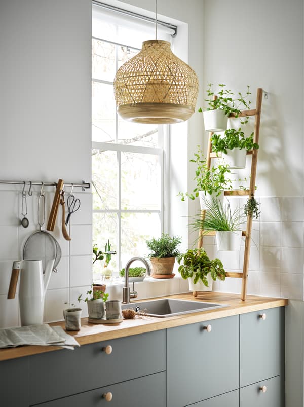 A kitchen with a ladder-like SATSUMAS bamboo plant stand with five plant pots standing on the worktop in front of a window.