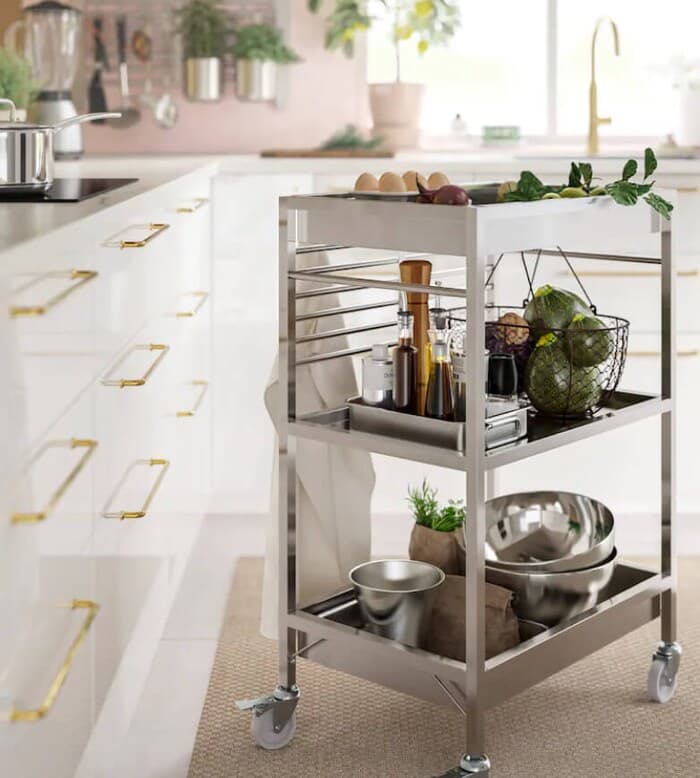 A kitchen with a pink wall has wall racks holding utensils and white drawers and cabinets with brass coloured details.
