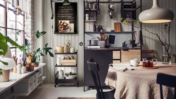 A kitchen with an ÄSPINGE kitchenette with a NISSAFORS trolley in black holding diverse items, a kitchen table with chairs.