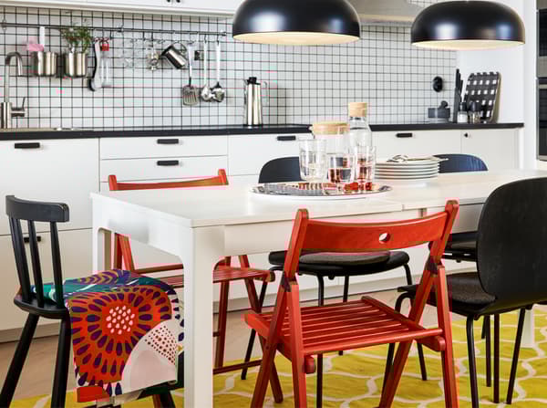 A kitchen with an extended table in white, red and black chairs, a yellow/white rug and two black pendant lamps.