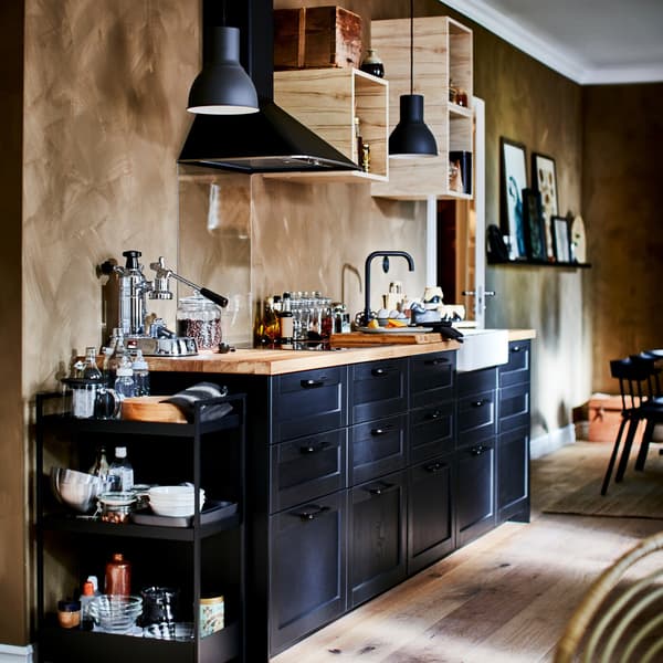A kitchen with black drawer fronts, a wooden worktop, a black trolley, open wall cabinets and two dark grey pendant lamps.