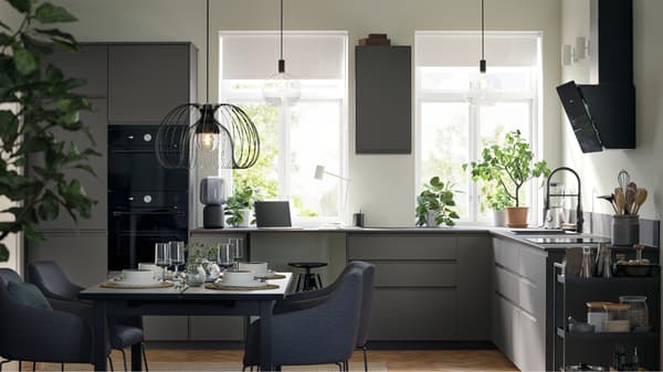 A kitchen with dark grey VOXTORP fronts and black appliances has three pendant lamps and a dining area with four chairs.