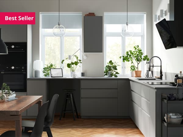 A kitchen with dark grey VOXTORP fronts and two big windows with a wall cabinet between them and green plants on the worktop.