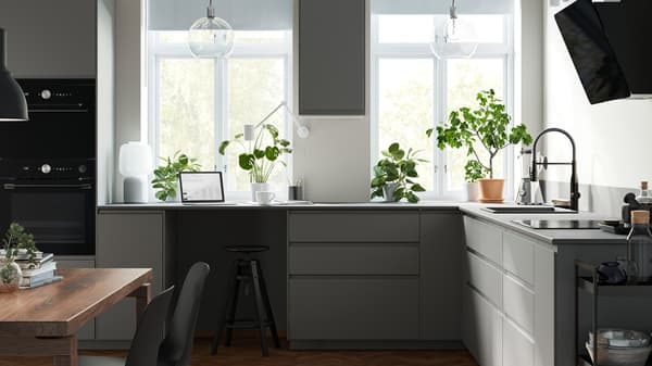 A kitchen with dark grey VOXTORP fronts and two big windows with a wall cabinet between them and green plants on the worktop.