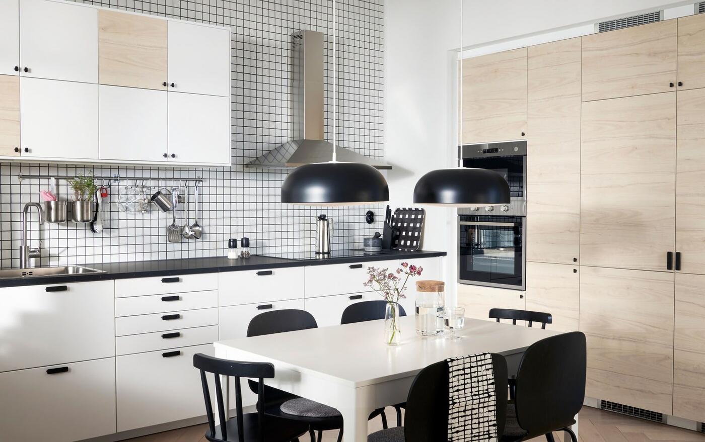 A kitchen with doors in white and light ash effect, two black pendant lamps, a white table and black chairs.