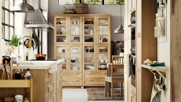 A kitchen with FORSBACKA fronts in oak veneer, including a glass-door cabinet with various kitchen items in it.
