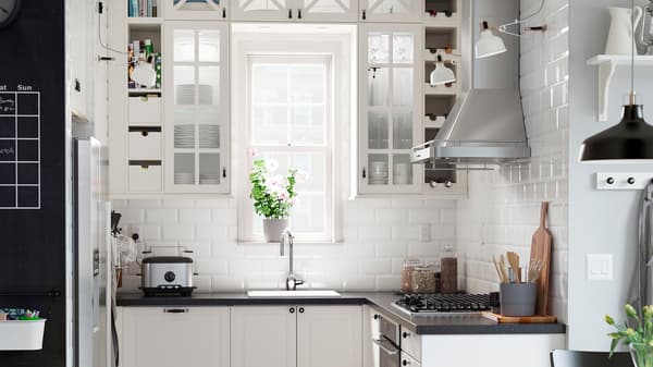 A kitchen with off-white glass-doored cabinets arranged around a window and a black mineral effect worktop.