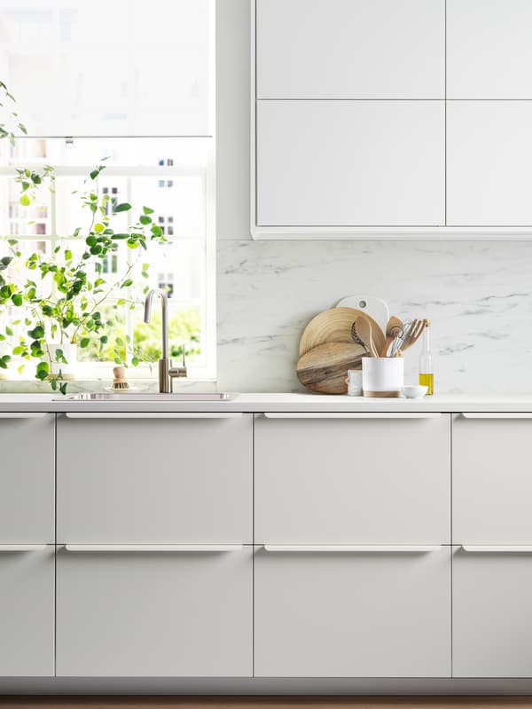 A kitchen with white cabinet fronts, a white worktop, a white marble-effect/quartz splashback and a plant next to a window.