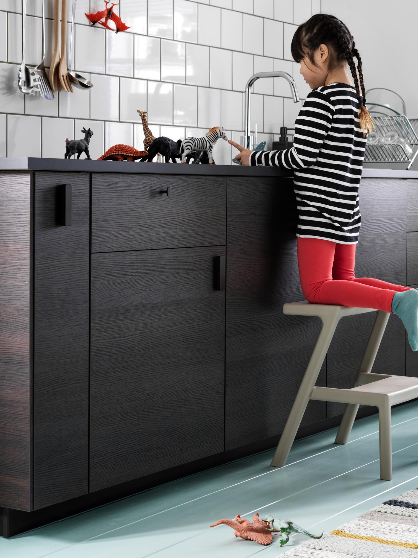 A kitchen with white-tiled walls and ASKERSUND drawers in dark brown. A young girl is playing with toys on the worktop.