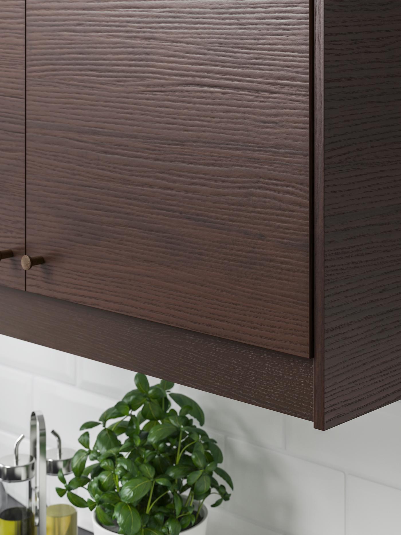 A kitchen with white tiles, a wall mounted ASKERSUND cabinet in dark drown ash effect, a pot of basil is underneath.