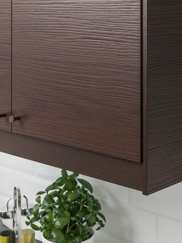 A kitchen with white tiles, a wall mounted ASKERSUND cabinet in dark drown ash effect, a pot of basil is underneath.