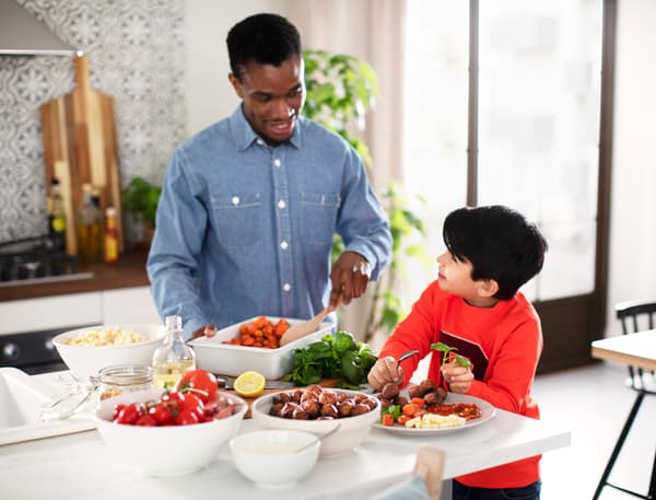A kitchen worktop with bowls of cooked carrots, pasta, plant balls and tomatoes. An adult serves food to a child.