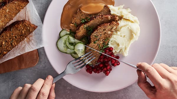 A knife and fork about to dig into a plate of meatloaf, sliced cucumbers, lingonberries, cream sauce and mashed potatoes.