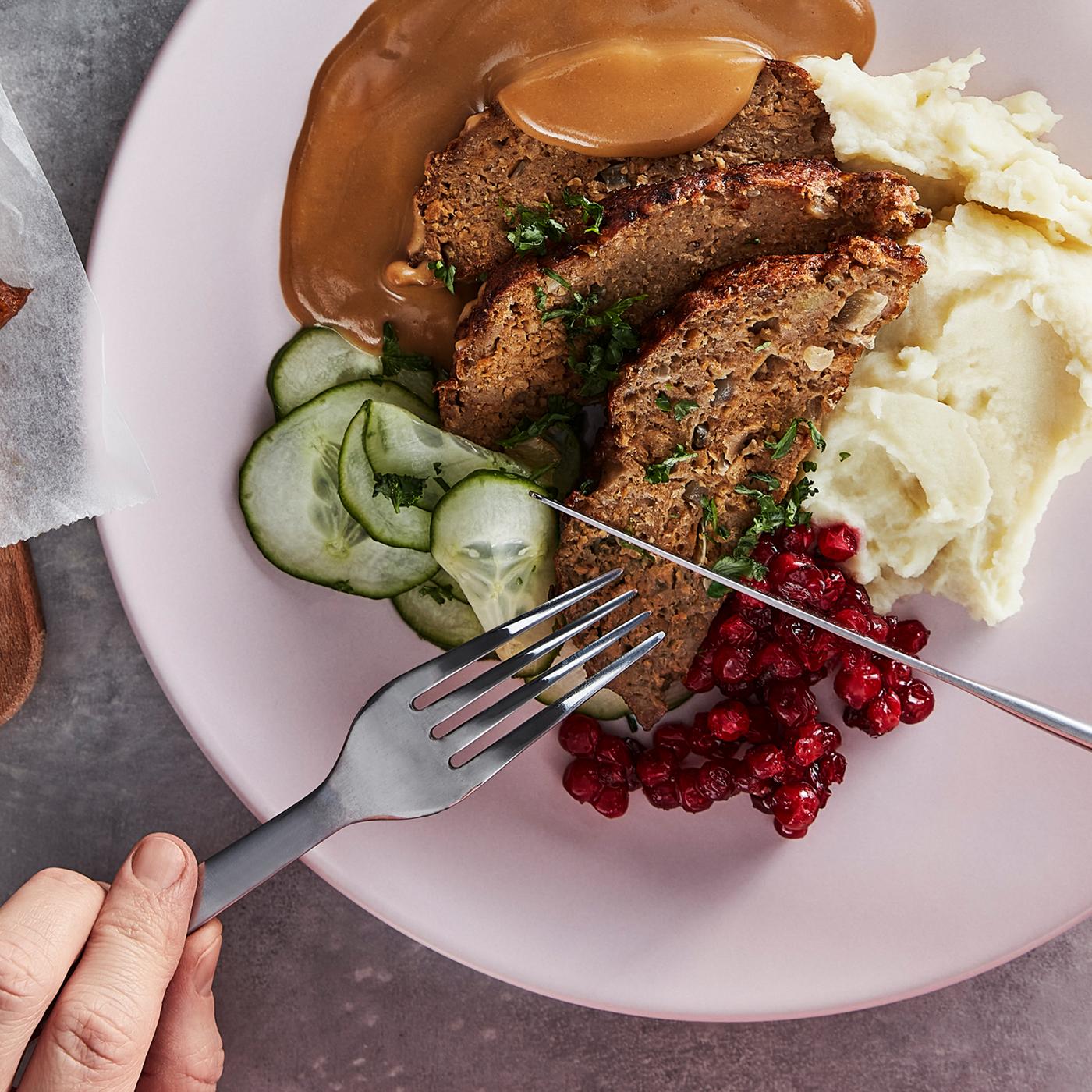 A knife and fork, held above a plate of plant-based mince loaf, cucumbers, lingonberries, cream sauce and mashed potatoes.
