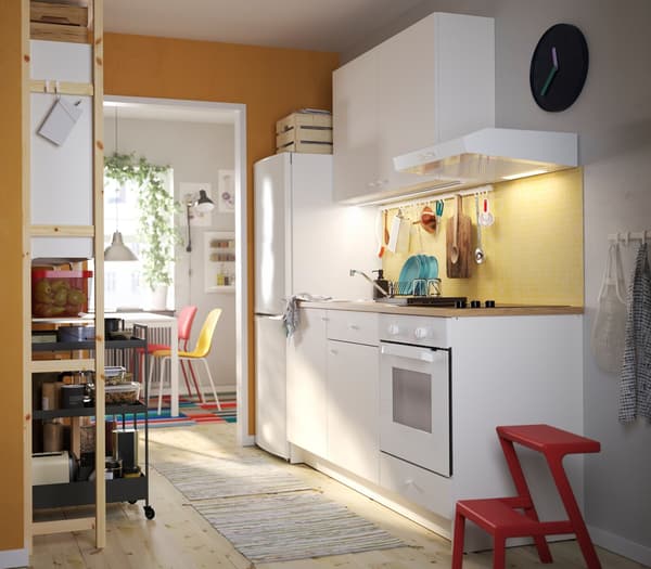 A KNOXHULT kitchen in white, a red step stool, a shelving unit in pine, black trolleys and colourful kitchen chairs.