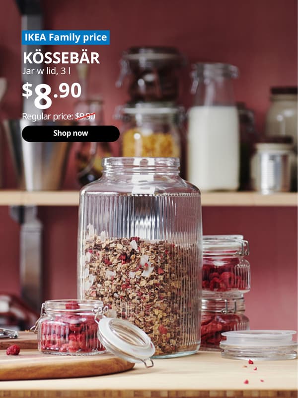 A KÖSSEBÄR jar with lid that is filled with granola placed on a RIMFORSA work bench in a kitchen setting.  Blue tag with white text that reads "IKEA Family price". Followed by white text over image that reads "KÖSSEBÄR, Jar w lid, 3 l, Price $8.90" Regular price: $9.90 with red line slashed across.