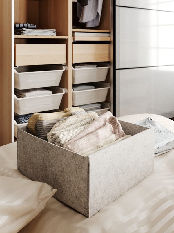 A KOMPLEMENT box on a bed, holding neatly folded garments, behind it is an open wardrobe with both drawers and hanging clothes.