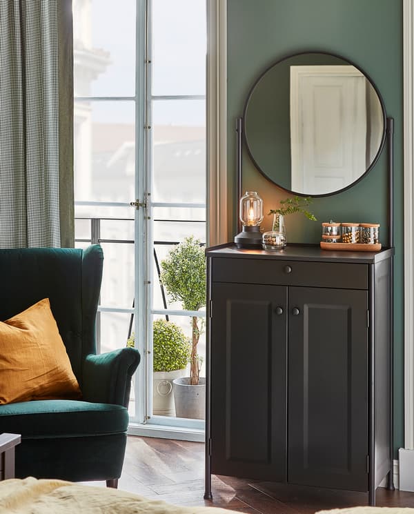 A KORNSJÖ cabinet with mirror stands by a wall. A table lamp, a vase and glass jars with lids stand on its top surface.