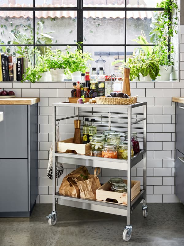 A KUNGSFORS kitchen trolley in stainless steel with three shelves and wheels, filled with spices and pickle jars.