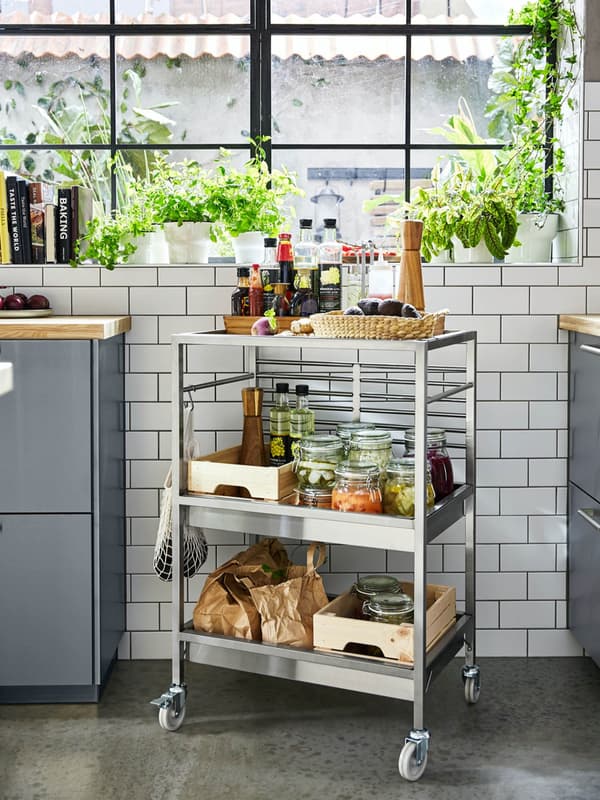 A KUNGSFORS kitchen trolley in stainless steel with three shelves and wheels, filled with spices and pickle jars.