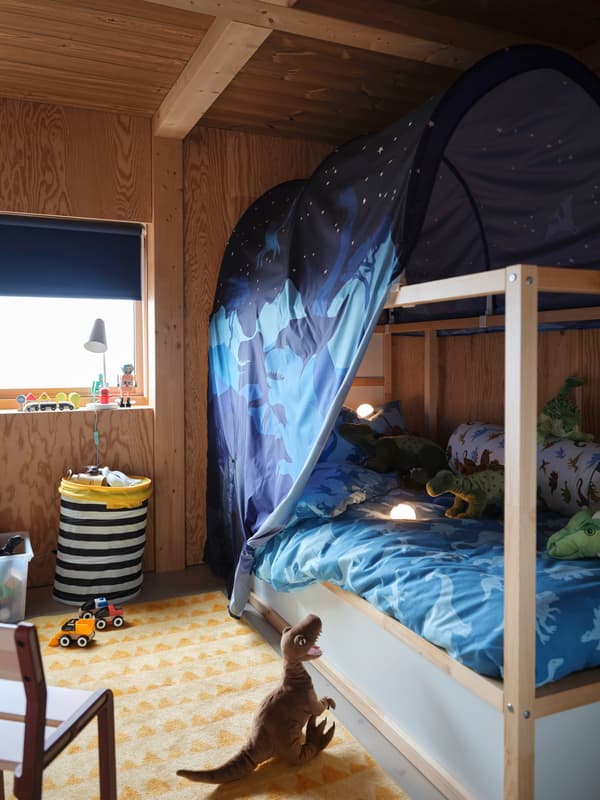 A KURA bed with dinosaur-print bed tent and duvet is covered with dinosaur soft toys, while another sits on the floor.