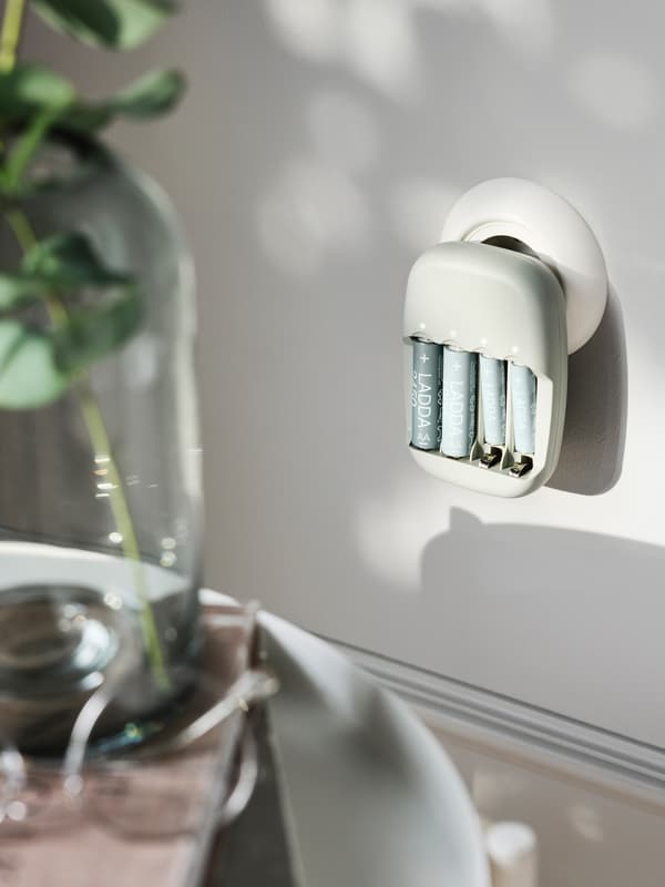 A LADDA battery charger charging batteries while plugged into a round power outlet in a light grey wall.