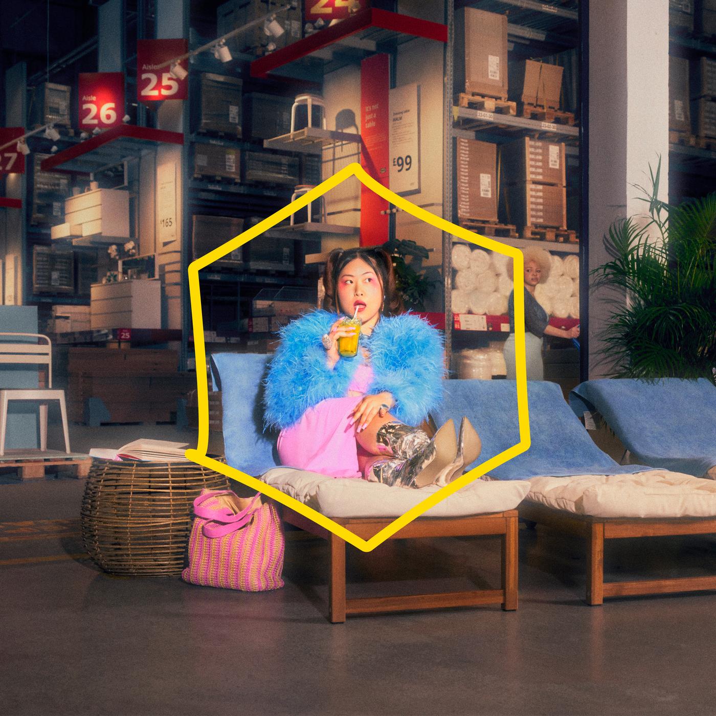 A lady sitting on an outdoor light brown stained NÄMMARÖ sun lounger in an IKEA store warehouse