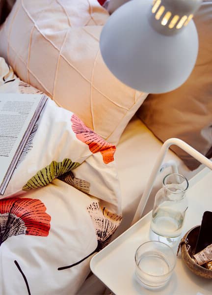 A lamp shines on floral bedding, pillows and a book, and a glass of water and carafe on a small white side table.